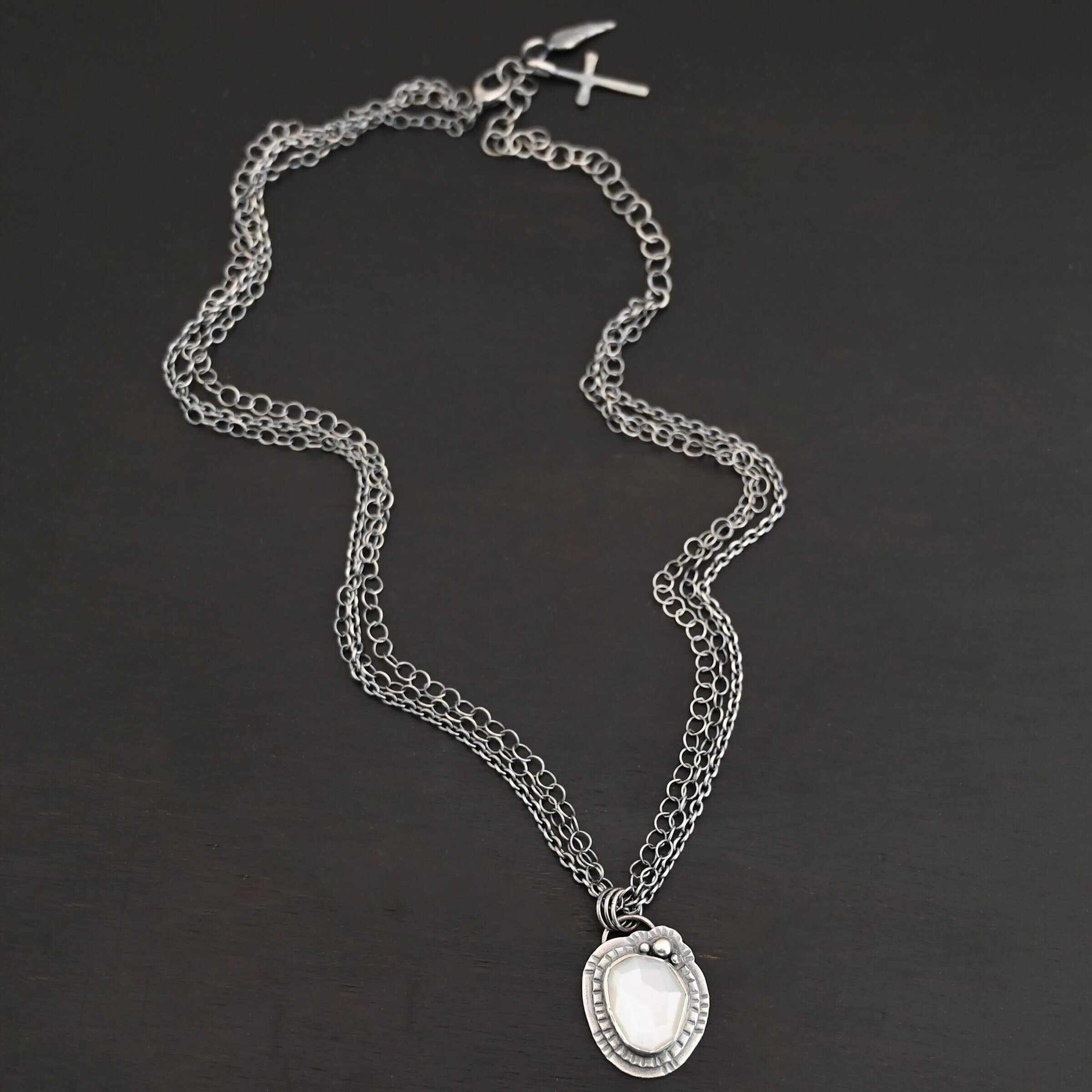 Inara Necklace "Inara" Necklace. There is an oval, vertical moonstone in the center. There are three balls of silver on the top right side. There are two layers of hammered sterling silver around the rest of the stone. The chain of the necklace is multiple chains that meet at the back.
