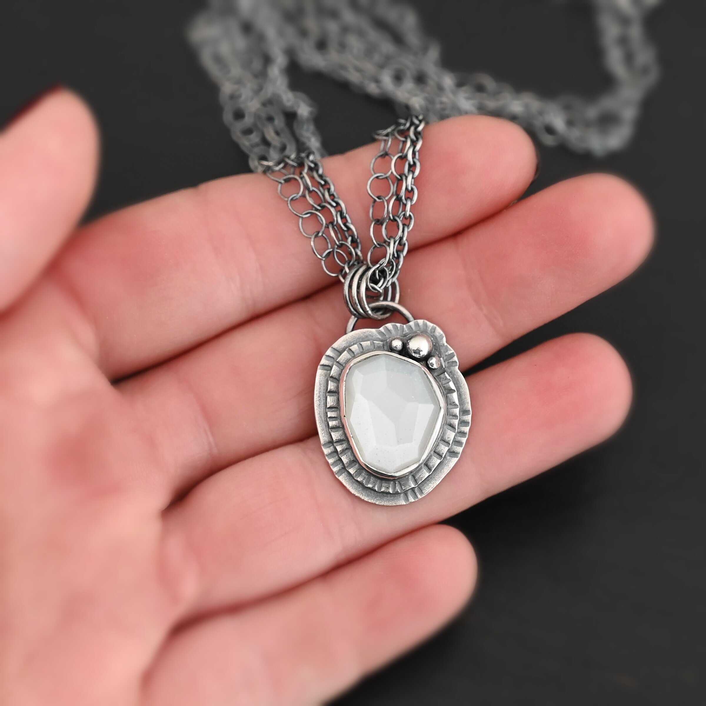 Inara Necklace "Inara" Necklace. There is an oval, vertical moonstone in the center. There are three balls of silver on the top right side. There are two layers of hammered sterling silver around the rest of the stone. The chain of the necklace is multiple chains that meet at the back. Necklace is held in the fingertips of a hand.