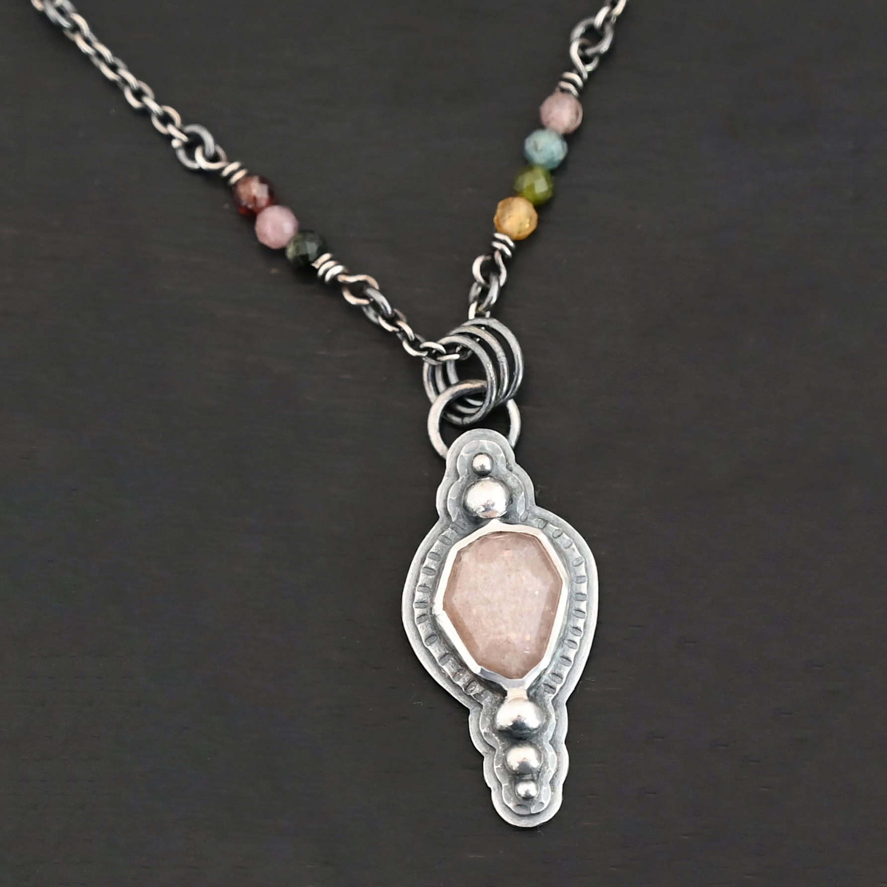 Leo Necklace "Leo" Necklace. There is an oval peach moonstone in the center. There are two balls of silver above it and three below it. The rest of the silver around the stone is hammered. There is a larger layer behind that which is smooth. There is a loop soldered to the back with three jump rings attached to it. The chain goes through them, and the chain has small tourmaline beads.
