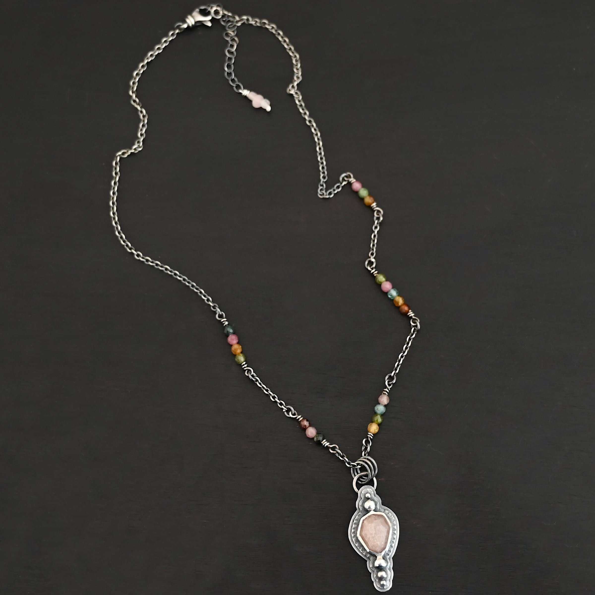 Leo Necklace "Leo" Necklace. There is an oval peach moonstone in the center. There are two balls of silver above it and three below it. The rest of the silver around the stone is hammered. There is a larger layer behind that which is smooth. There is a loop soldered to the back with three jump rings attached to it. The chain goes through them, and the chain has small tourmaline beads.