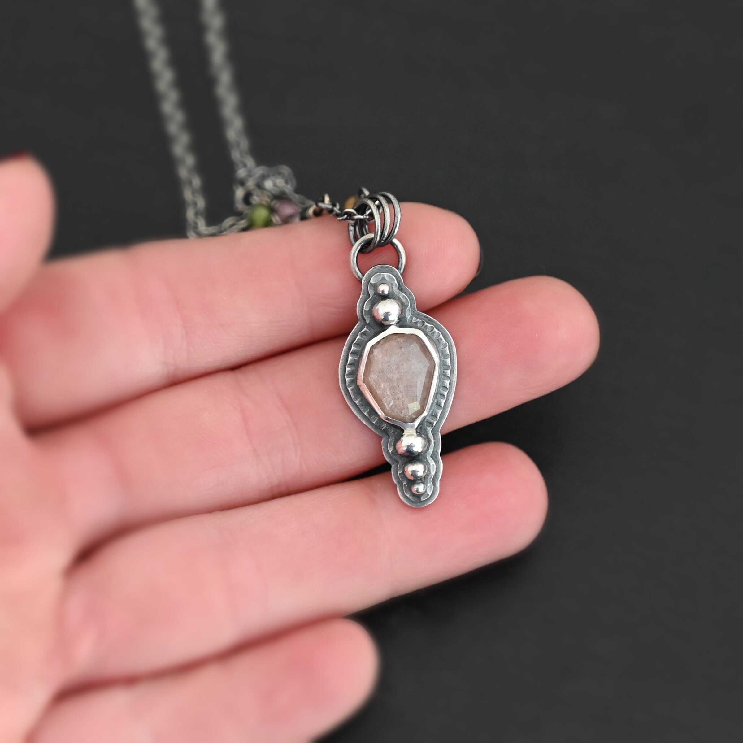 Leo Necklace "Leo" Necklace. There is an oval peach moonstone in the center. There are two balls of silver above it and three below it. The rest of the silver around the stone is hammered. There is a larger layer behind that which is smooth. There is a loop soldered to the back with three jump rings attached to it. The chain goes through them, and the chain has small tourmaline beads. Necklace is held in the fingertips of a hand.