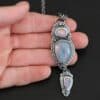 "Blue Moon Magic" Necklace. There are three opals in a line down. The top one is a small horizontal oval. The next one is a larger, vertical opal. The bottom one is a small triangle. Each one has silver ball detail surrounding it, as well as hammered silver. Necklace is lying in the fingertips of a hand.