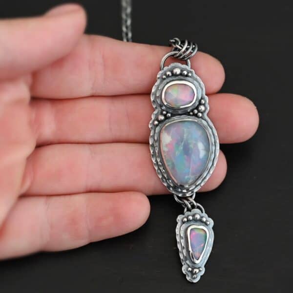 "Blue Moon Magic" Necklace. There are three opals in a line down. The top one is a small horizontal oval. The next one is a larger, vertical opal. The bottom one is a small triangle. Each one has silver ball detail surrounding it, as well as hammered silver. Necklace is lying in the fingertips of a hand.