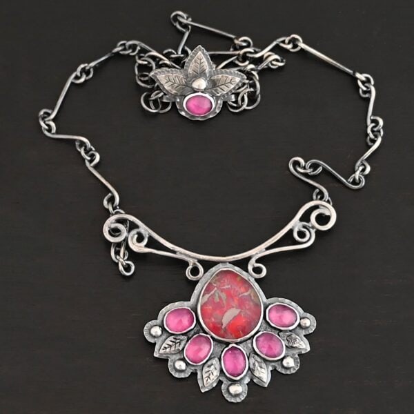 "In The Dawns" Necklace. There are six rubies and one large, wide, pear shaped red/pink opal pointing up. The opal is at the bottom, with five oval vertical rubies circling around the bottom of it. In between each ruby is a silver leaf and on the end of each ruby is a ball of silver. The clasp of the necklace has three silver leaves and a ball of silver above an oval ruby. Chain is handmade out of wavy links of sterling silver.