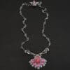 "In The Dawns" Necklace. There are six rubies and one large, wide, pear shaped red/pink opal pointing up. The opal is at the bottom, with five oval vertical rubies circling around the bottom of it. In between each ruby is a silver leaf and on the end of each ruby is a ball of silver. The clasp of the necklace has three silver leaves and a ball of silver above an oval ruby. Chain is handmade out of wavy links of sterling silver.