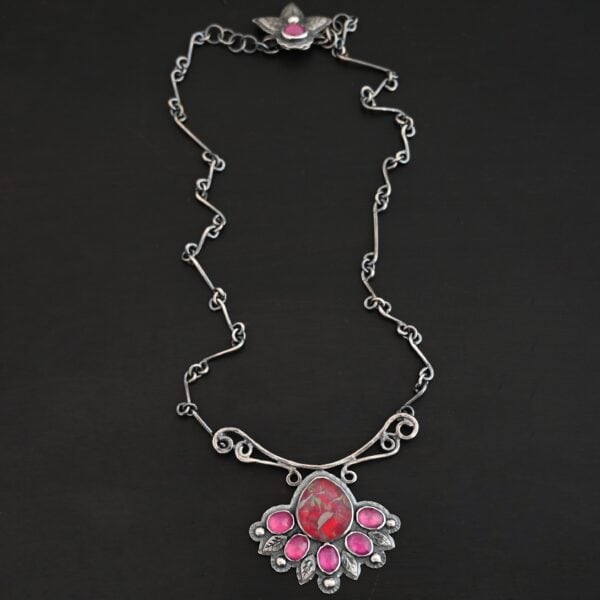 "In The Dawns" Necklace. There are six rubies and one large, wide, pear shaped red/pink opal pointing up. The opal is at the bottom, with five oval vertical rubies circling around the bottom of it. In between each ruby is a silver leaf and on the end of each ruby is a ball of silver. The clasp of the necklace has three silver leaves and a ball of silver above an oval ruby. Chain is handmade out of wavy links of sterling silver.