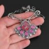 "In The Dawns" Necklace. There are six rubies and one large, wide, pear shaped red/pink opal pointing up. The opal is at the bottom, with five oval vertical rubies circling around the bottom of it. In between each ruby is a silver leaf and on the end of each ruby is a ball of silver. The clasp of the necklace has three silver leaves and a ball of silver above an oval ruby. Chain is handmade out of wavy links of sterling silver. Necklace is lying in the fingertips of a hand.