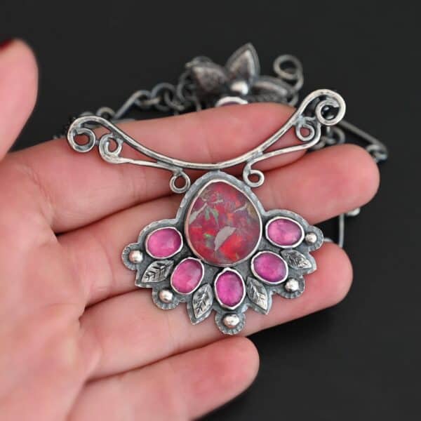 "In The Dawns" Necklace. There are six rubies and one large, wide, pear shaped red/pink opal pointing up. The opal is at the bottom, with five oval vertical rubies circling around the bottom of it. In between each ruby is a silver leaf and on the end of each ruby is a ball of silver. The clasp of the necklace has three silver leaves and a ball of silver above an oval ruby. Chain is handmade out of wavy links of sterling silver. Necklace is lying in the fingertips of a hand.