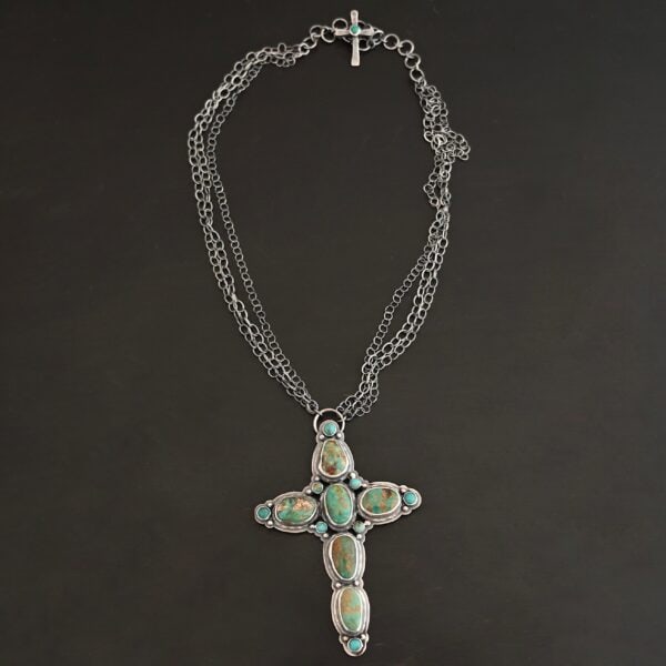 "Firm In Faith" Necklace. Necklace is a large cross shape with six oval turquoise creating the cross shape and eight small circular turquoise around the rest of the pendant. Chain is multiple strands that meet at the back. Necklace has a small silver cross with a turquoise in the center of it for the clasp.