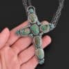 "Firm In Faith" Necklace. Necklace is a large cross shape with six oval turquoise creating the cross shape and eight small circular turquoise around the rest of the pendant. Chain is multiple strands that meet at the back. Necklace is lying in the fingertips of a hand.