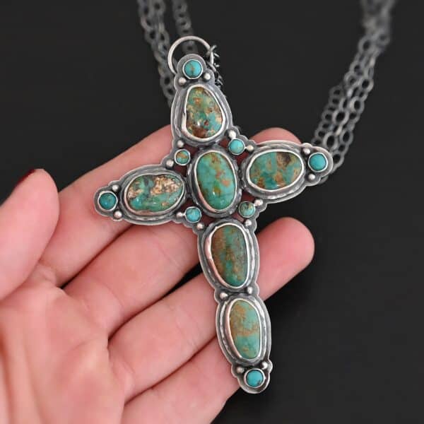 "Firm In Faith" Necklace. Necklace is a large cross shape with six oval turquoise creating the cross shape and eight small circular turquoise around the rest of the pendant. Chain is multiple strands that meet at the back. Necklace is lying in the fingertips of a hand.