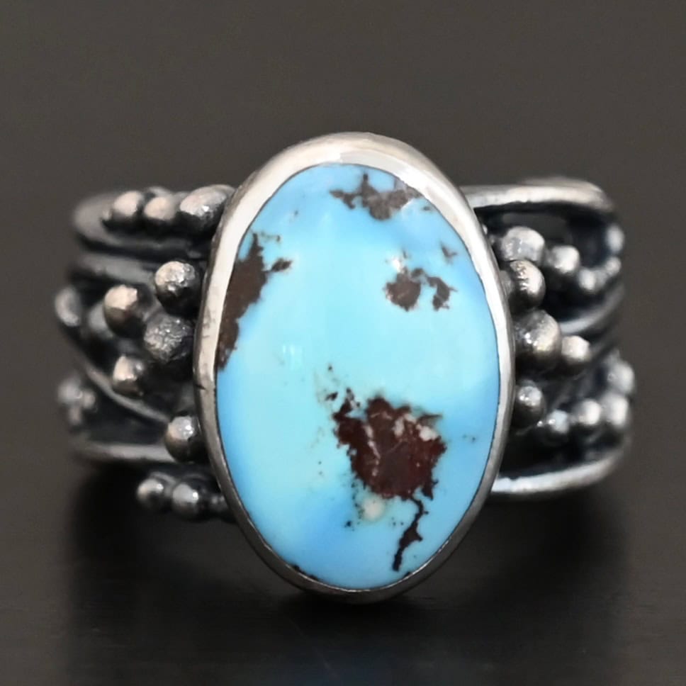 Morning Breeze Ring "Morning Breeze" Ring. There is an oval vertical turquoise in the center. The ring band is wide, with multiple lines of silver that are wavy and leave small gaps in the band. There are multiple balls of silver around the ring band.