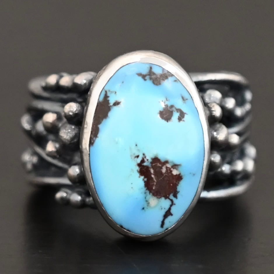 Morning Breeze Ring "Morning Breeze" Ring. There is an oval vertical turquoise in the center. The ring band is wide, with multiple lines of silver that are wavy and leave small gaps in the band. There are multiple balls of silver around the ring band.