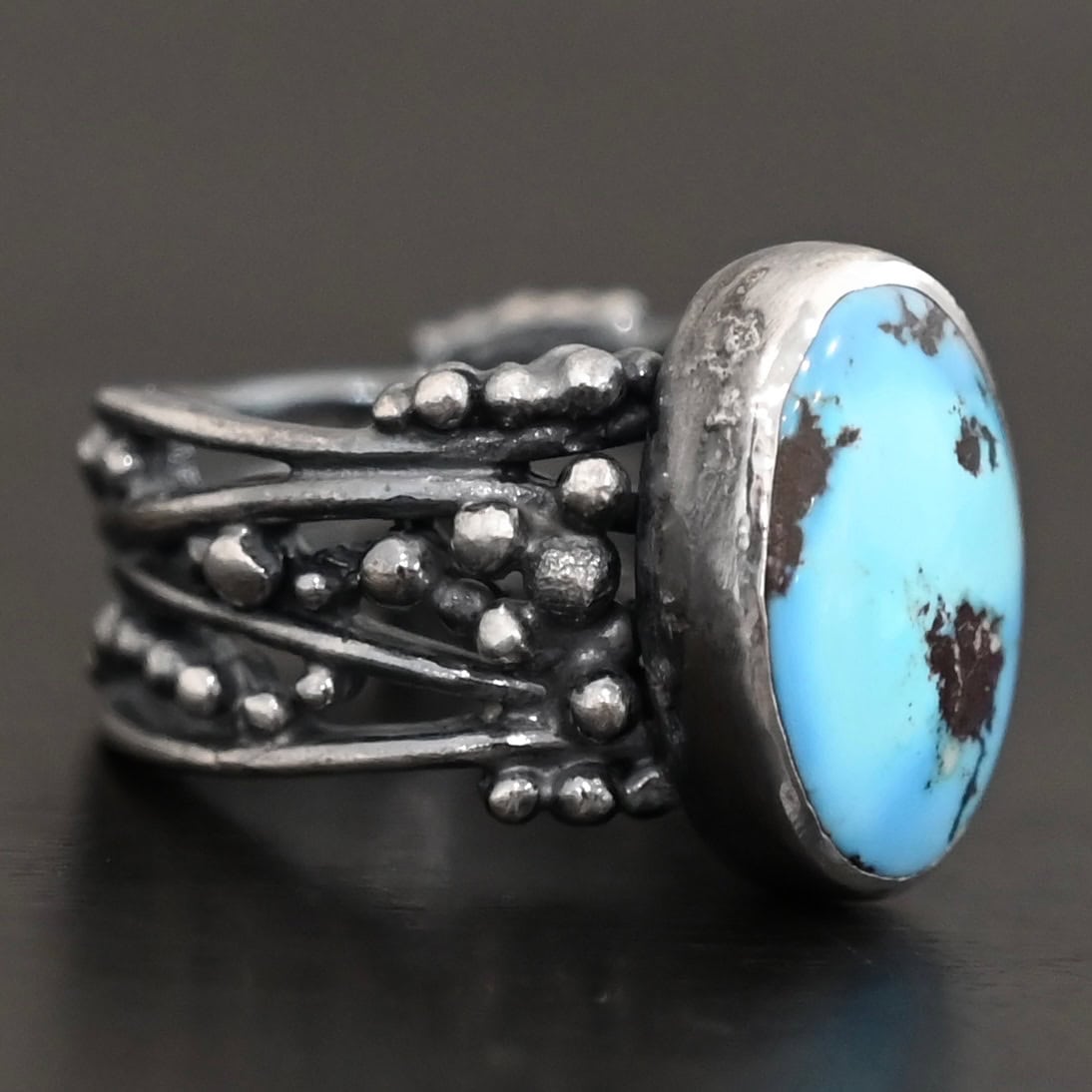 Morning Breeze Ring "Morning Breeze" Ring. There is an oval vertical turquoise in the center. The ring band is wide, with multiple lines of silver that are wavy and leave small gaps in the band. There are multiple balls of silver around the ring band.
