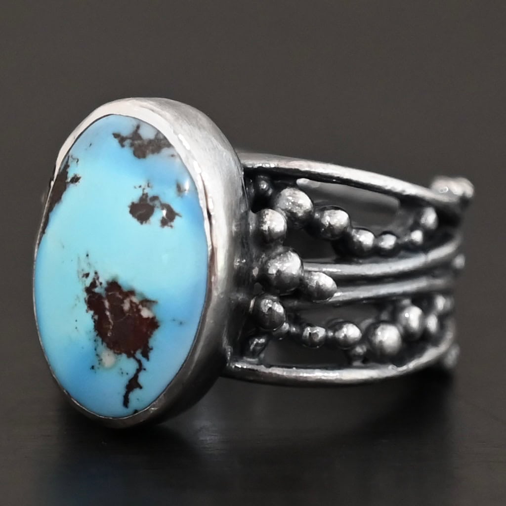Morning Breeze Ring "Morning Breeze" Ring. There is an oval vertical turquoise in the center. The ring band is wide, with multiple lines of silver that are wavy and leave small gaps in the band. There are multiple balls of silver around the ring band.