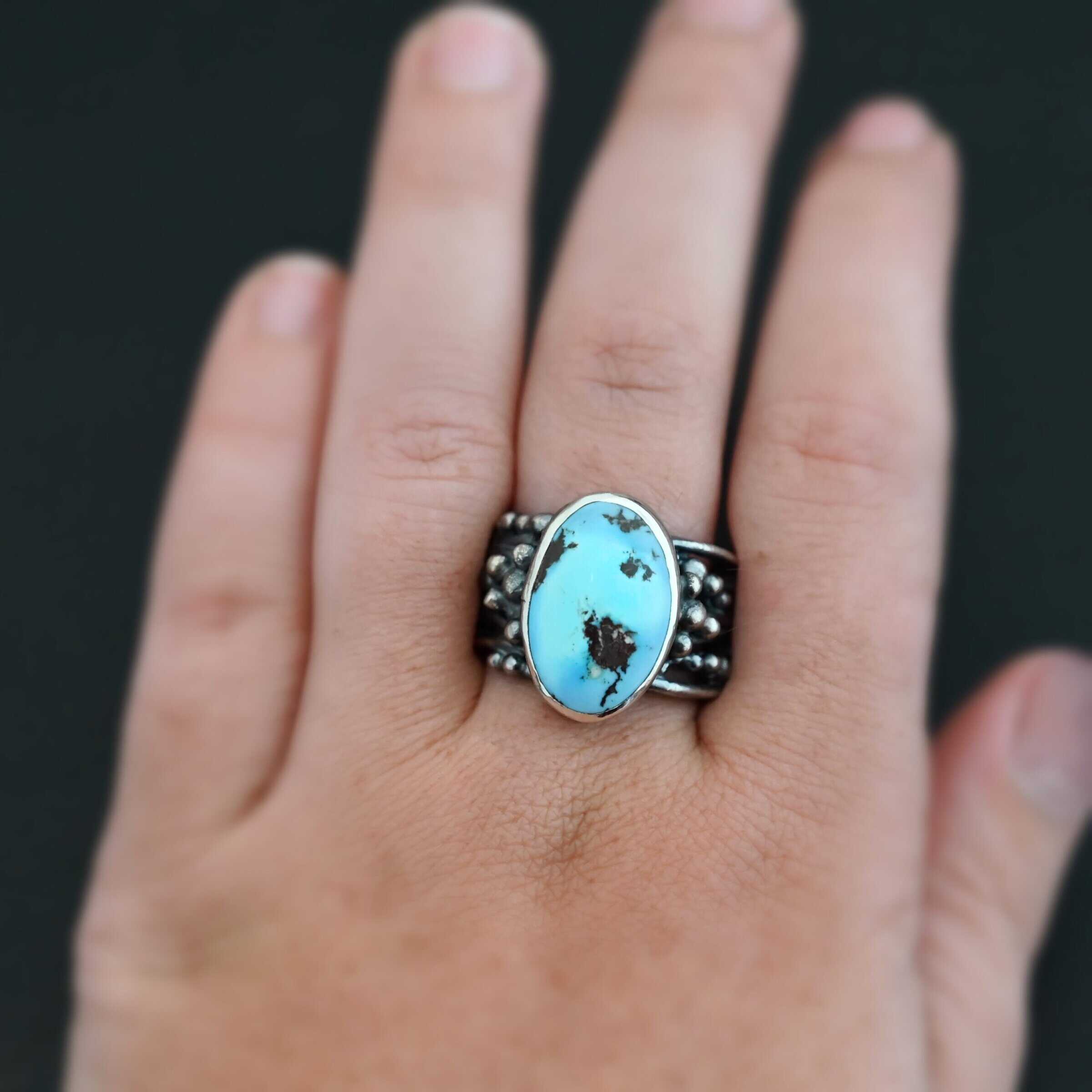 Morning Breeze Ring "Morning Breeze" Ring. There is an oval vertical turquoise in the center. The ring band is wide, with multiple lines of silver that are wavy and leave small gaps in the band. There are multiple balls of silver around the ring band. Ring is worn up close by a model.