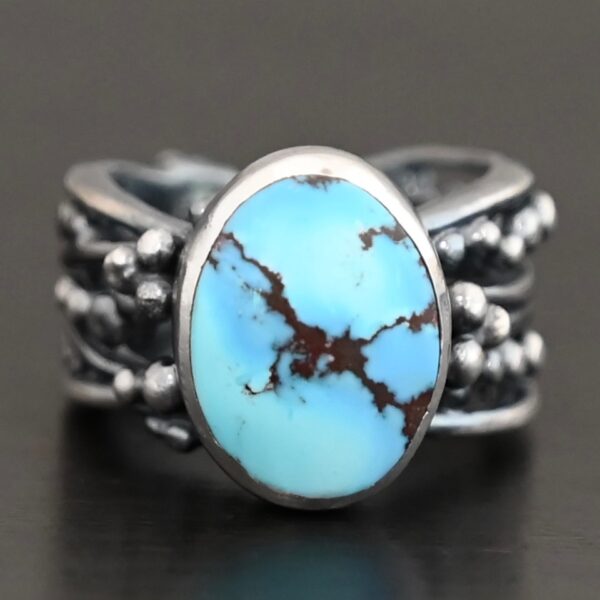 "Over The Horizon" Ring. There is an oval vertical turquoise in the center. The ring band is wide, with multiple lines of silver that are wavy and leave small gaps in the band. There are multiple balls of silver around the ring band.
