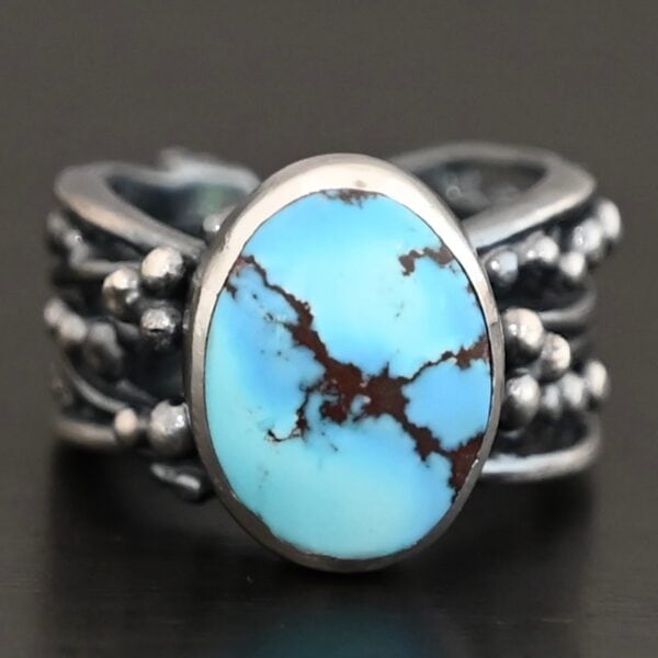 "Over The Horizon" Ring. There is an oval vertical turquoise in the center. The ring band is wide, with multiple lines of silver that are wavy and leave small gaps in the band. There are multiple balls of silver around the ring band.
