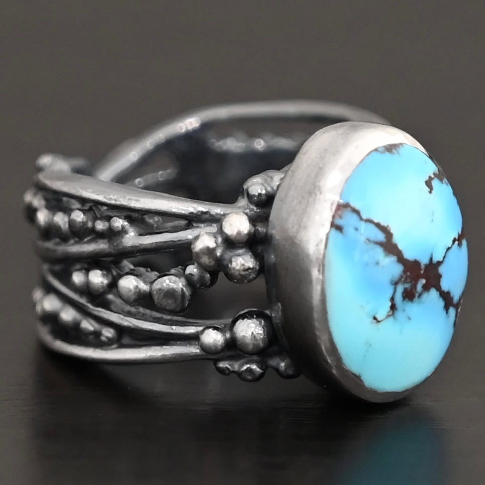 Over The Horizon Ring "Over The Horizon" Ring. There is an oval vertical turquoise in the center. The ring band is wide, with multiple lines of silver that are wavy and leave small gaps in the band. There are multiple balls of silver around the ring band.