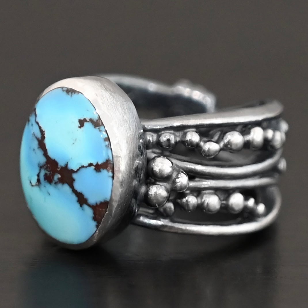 Over The Horizon Ring "Over The Horizon" Ring. There is an oval vertical turquoise in the center. The ring band is wide, with multiple lines of silver that are wavy and leave small gaps in the band. There are multiple balls of silver around the ring band.