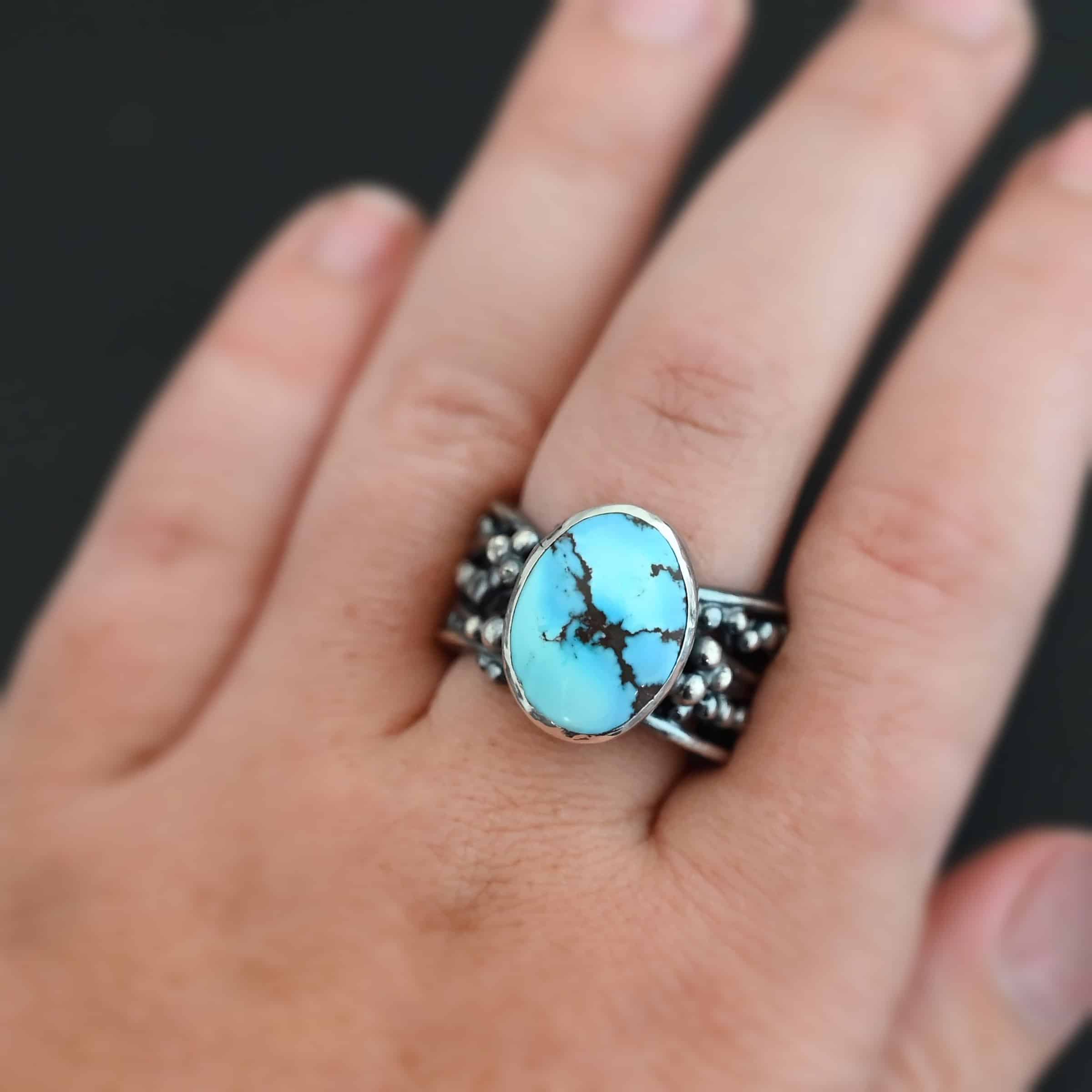 Over The Horizon Ring "Over The Horizon" Ring. There is an oval vertical turquoise in the center. The ring band is wide, with multiple lines of silver that are wavy and leave small gaps in the band. There are multiple balls of silver around the ring band. Ring is worn up close by a model.