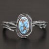 "Grander Than The Sky" Cuff. There is an oval turquoise in the center, vertical. There are two lines of hammered silver that wrap around the stone and each other. There is a gap in between the lines of silver and turquoise that is oxidized. The cuff is casted sterling silver branches from a tree that are curved.