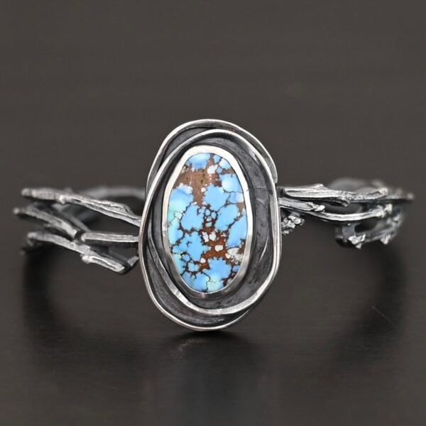 "Grander Than The Sky" Cuff. There is an oval turquoise in the center, vertical. There are two lines of hammered silver that wrap around the stone and each other. There is a gap in between the lines of silver and turquoise that is oxidized. The cuff is casted sterling silver branches from a tree that are curved.