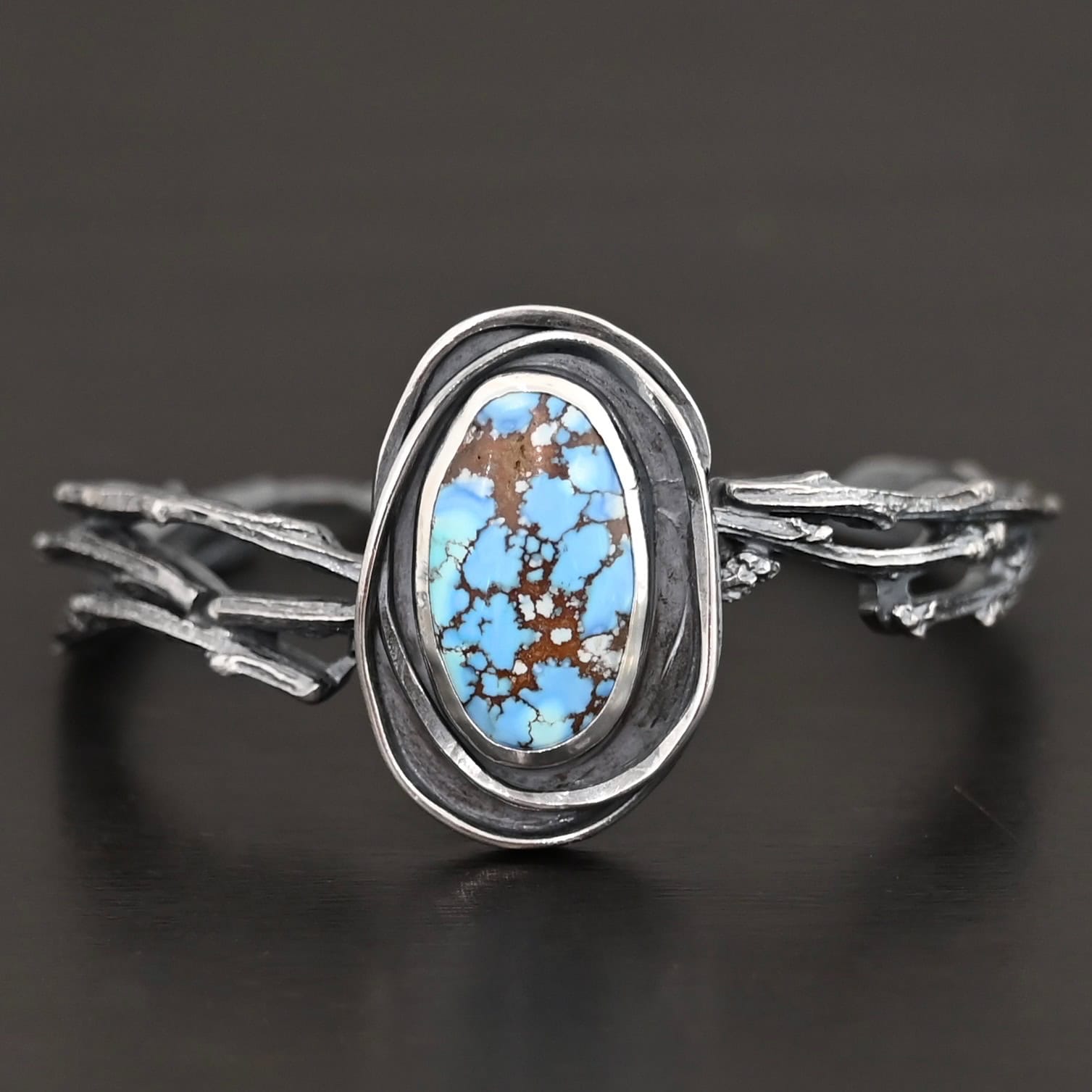 "Loire" Cuff. There is an oval turquoise in the center, vertical. There are two lines of hammered silver that wrap around the stone and each other. There is a gap in between the lines of silver and turquoise that is oxidized. The cuff is casted sterling silver branches from a tree that are curved.