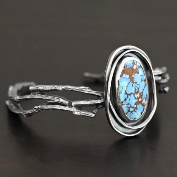 "Grander Than The Sky" Cuff. There is an oval turquoise in the center, vertical. There are two lines of hammered silver that wrap around the stone and each other. There is a gap in between the lines of silver and turquoise that is oxidized. The cuff is casted sterling silver branches from a tree that are curved.