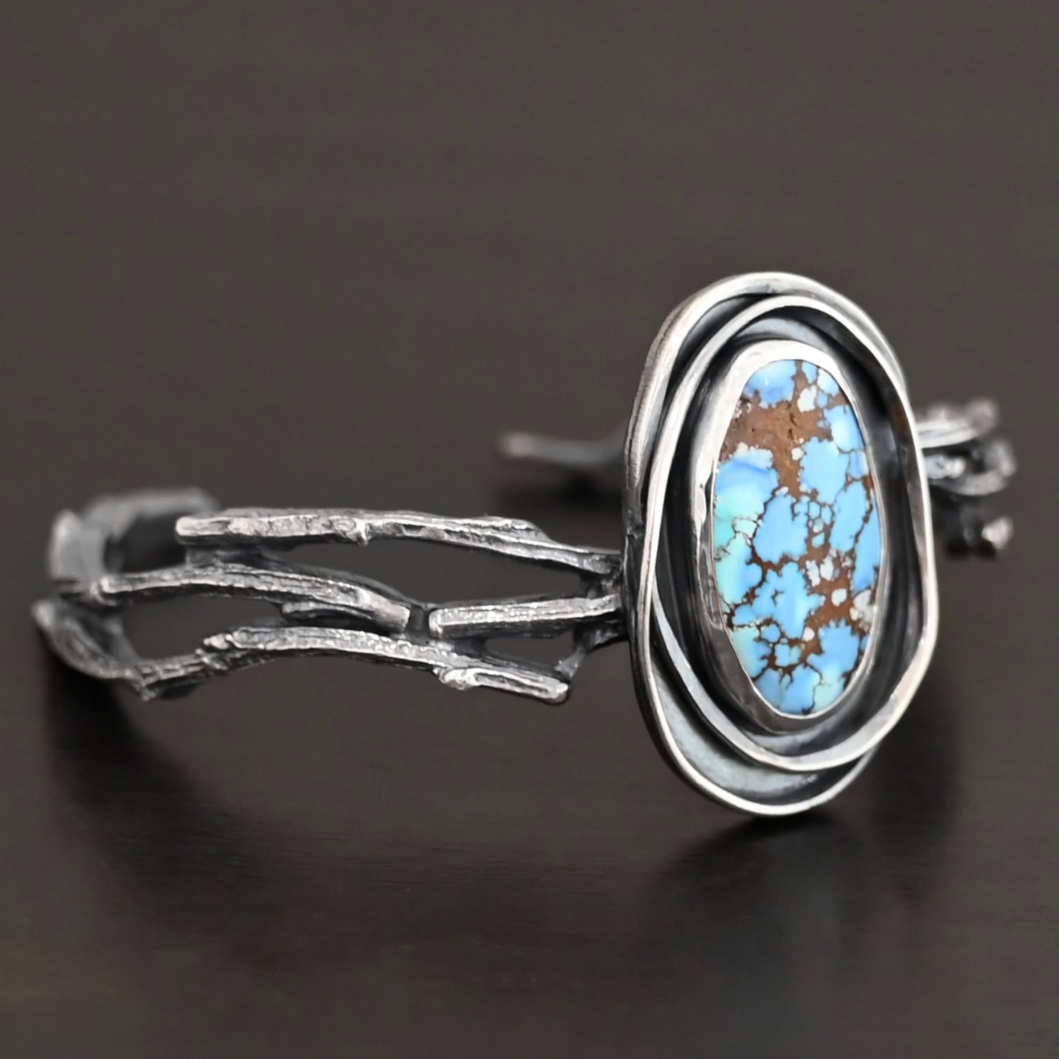 "Loire" Cuff. There is an oval turquoise in the center, vertical. There are two lines of hammered silver that wrap around the stone and each other. There is a gap in between the lines of silver and turquoise that is oxidized. The cuff is casted sterling silver branches from a tree that are curved.
