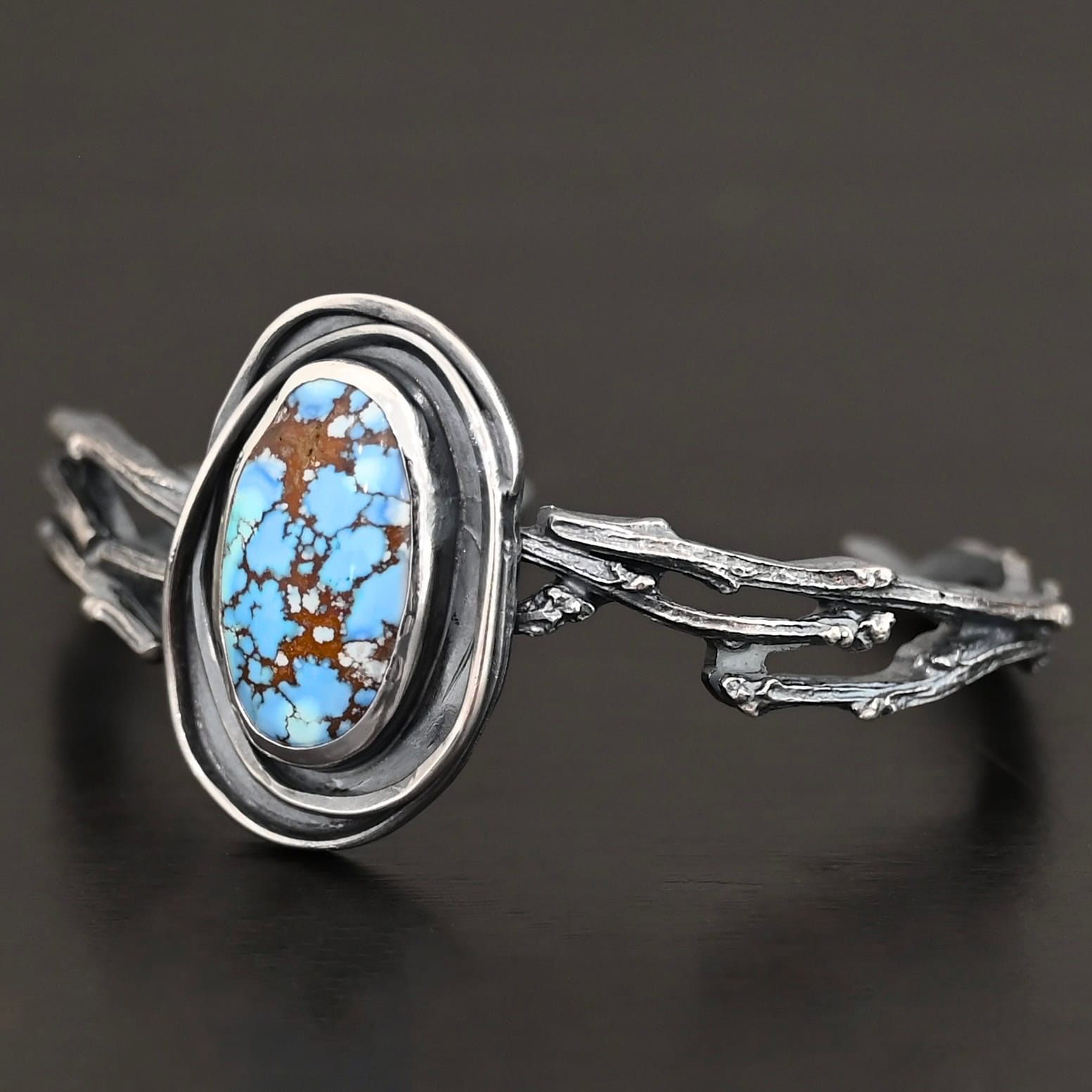 "Loire" Cuff. There is an oval turquoise in the center, vertical. There are two lines of hammered silver that wrap around the stone and each other. There is a gap in between the lines of silver and turquoise that is oxidized. The cuff is casted sterling silver branches from a tree that are curved.