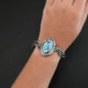 "Grander Than The Sky" Cuff. There is an oval turquoise in the center, vertical. There are two lines of hammered silver that wrap around the stone and each other. There is a gap in between the lines of silver and turquoise that is oxidized. The cuff is casted sterling silver branches from a tree that are curved. Cuff is worn up close by a model.