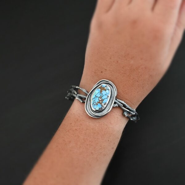"Grander Than The Sky" Cuff. There is an oval turquoise in the center, vertical. There are two lines of hammered silver that wrap around the stone and each other. There is a gap in between the lines of silver and turquoise that is oxidized. The cuff is casted sterling silver branches from a tree that are curved. Cuff is worn up close by a model.