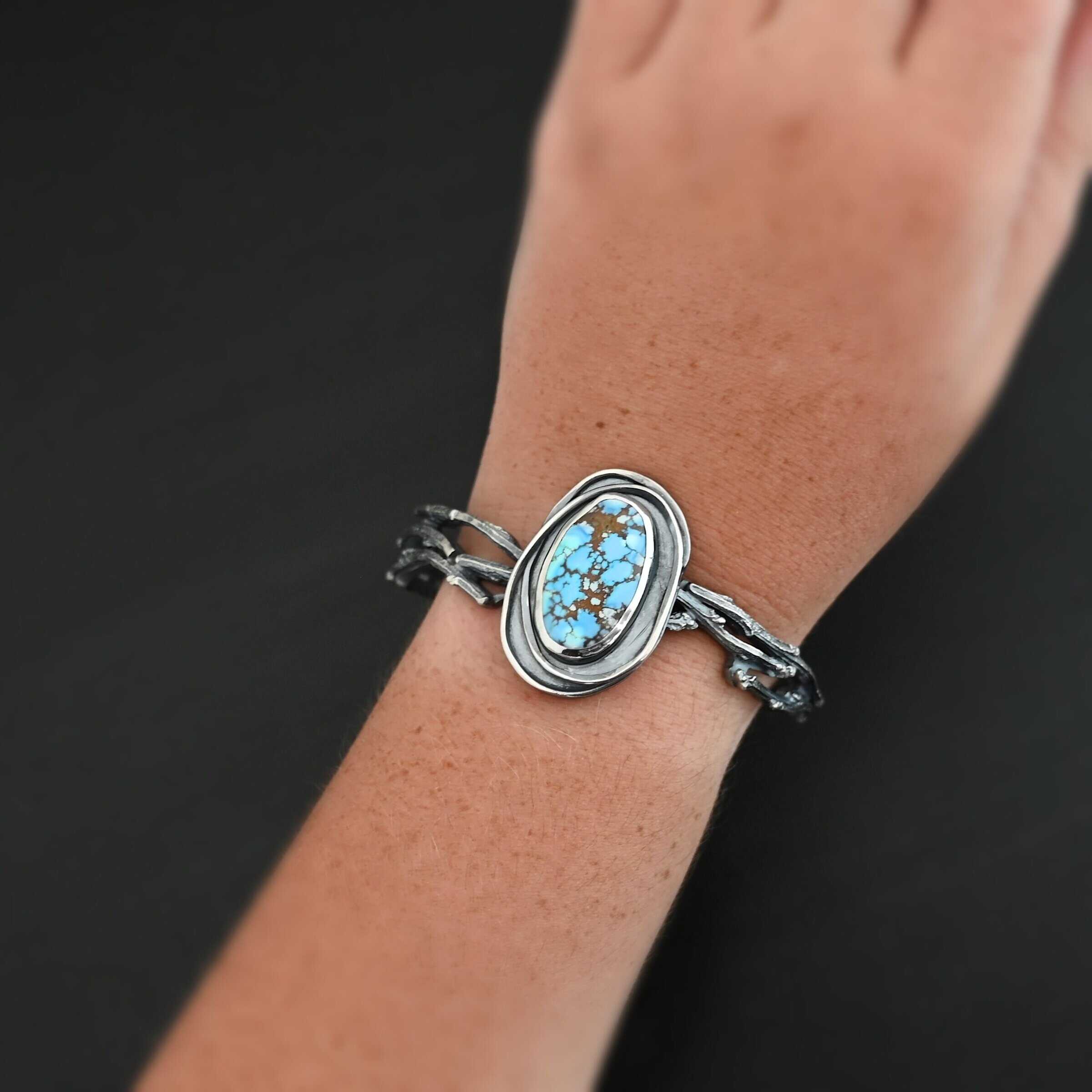"Loire" Cuff. There is an oval turquoise in the center, vertical. There are two lines of hammered silver that wrap around the stone and each other. There is a gap in between the lines of silver and turquoise that is oxidized. The cuff is casted sterling silver branches from a tree that are curved. Cuff is worn up close by a model.