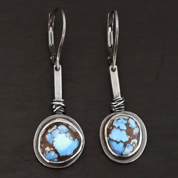 "Seven Summers Ago" Earrings. Each earring has a short line of vertical silver with silver wire wrapped around the bottom of it. Soldered to the bottom of the line is a circular turquoise with smooth silver around it. The wire is wrapped above the turquoise.