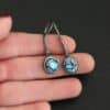 "Seven Summers Ago" Earrings. Each earring has a short line of vertical silver with silver wire wrapped around the bottom of it. Soldered to the bottom of the line is a circular turquoise with smooth silver around it. The wire is wrapped above the turquoise. Earrings are held in the fingertips of a hand.