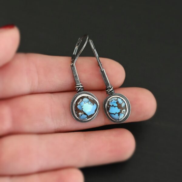 "Seven Summers Ago" Earrings. Each earring has a short line of vertical silver with silver wire wrapped around the bottom of it. Soldered to the bottom of the line is a circular turquoise with smooth silver around it. The wire is wrapped above the turquoise. Earrings are held in the fingertips of a hand.