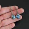 "Simmered In Sunshine" Earrings. Each earring has a short line of vertical silver with silver wire wrapped around the bottom of it. Soldered to the bottom of the line is a circular turquoise with smooth silver around it. The wire is wrapped above the turquoise. Earrings are held in the fingertips of a hand.