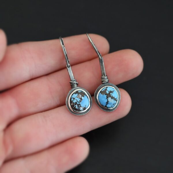 "Simmered In Sunshine" Earrings. Each earring has a short line of vertical silver with silver wire wrapped around the bottom of it. Soldered to the bottom of the line is a circular turquoise with smooth silver around it. The wire is wrapped above the turquoise. Earrings are held in the fingertips of a hand.