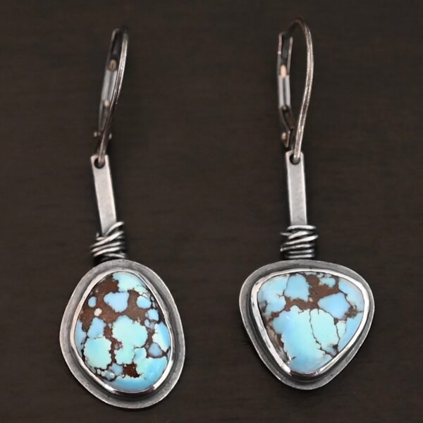 "Enzo" Earrings. Each earring has a short line of vertical silver with silver wire wrapped around the bottom of it. Soldered to the bottom of the line is an oval turquoise on one and a triangular on the other, both with smooth silver around them. The wire is wrapped above the turquoise.