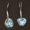 "Enzo" Earrings. Each earring has a short line of vertical silver with silver wire wrapped around the bottom of it. Soldered to the bottom of the line is an oval turquoise on one and a triangular on the other, both with smooth silver around them. The wire is wrapped above the turquoise.