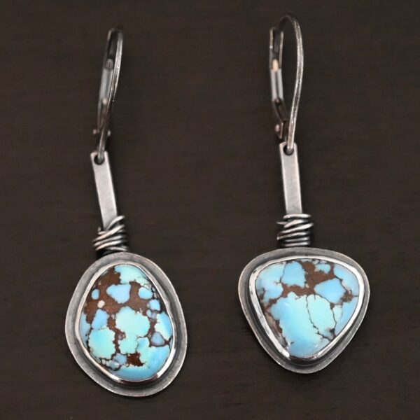 "Enzo" Earrings. Each earring has a short line of vertical silver with silver wire wrapped around the bottom of it. Soldered to the bottom of the line is an oval turquoise on one and a triangular on the other, both with smooth silver around them. The wire is wrapped above the turquoise.