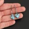 "Enzo" Earrings. Each earring has a short line of vertical silver with silver wire wrapped around the bottom of it. Soldered to the bottom of the line is an oval turquoise on one and a triangular on the other, both with smooth silver around them. The wire is wrapped above the turquoise. Earrings are held in the fingertips of a hand.