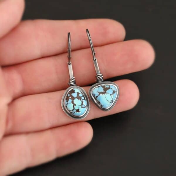 "Enzo" Earrings. Each earring has a short line of vertical silver with silver wire wrapped around the bottom of it. Soldered to the bottom of the line is an oval turquoise on one and a triangular on the other, both with smooth silver around them. The wire is wrapped above the turquoise. Earrings are held in the fingertips of a hand.