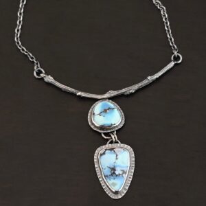 "Story To Tell" Necklace. There are two turquoise. The top one is a circular shape. The bottom one is a triangular shape. Both have hammered silver around them. The bottom stone hangs down from the top one. The top stone is soldered to a casted, curved sterling silver twig from a branch.