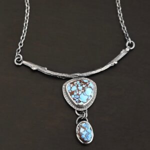 "New Beginnings" Necklace. There are two turquoise. The top one is a triangular shape. The bottom one is an oval. The top stone has hammered silver around it. The bottom stone hangs down from the top one. The top stone is soldered to a casted, curved sterling silver twig from a branch.