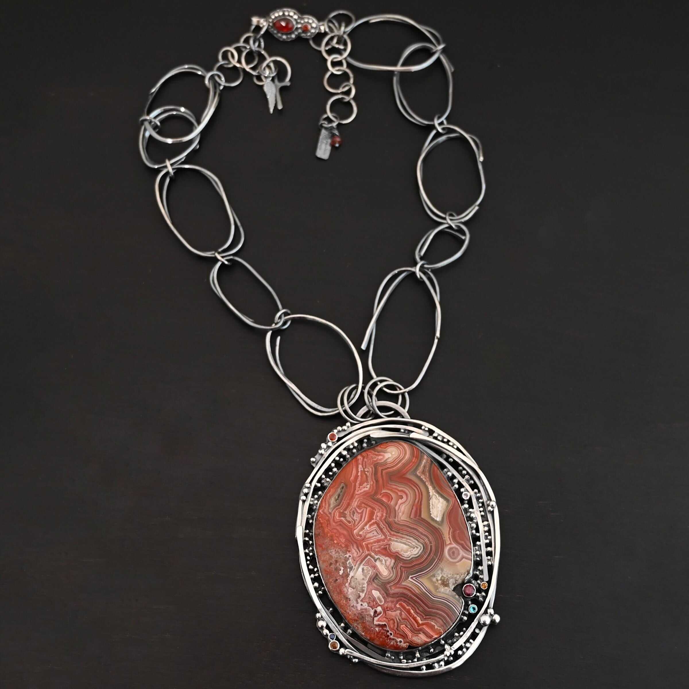 "Back To Our Roots" Necklace. There is a very large oval crazy lace agate in the center with multiple lines of sterling silver wrapped around it. There are small stones scattered around the pendant, along with lots of sterling silver ball detail. Chain is handmade out of large, sterling silver oval links that detach from the pendant to change the chain.