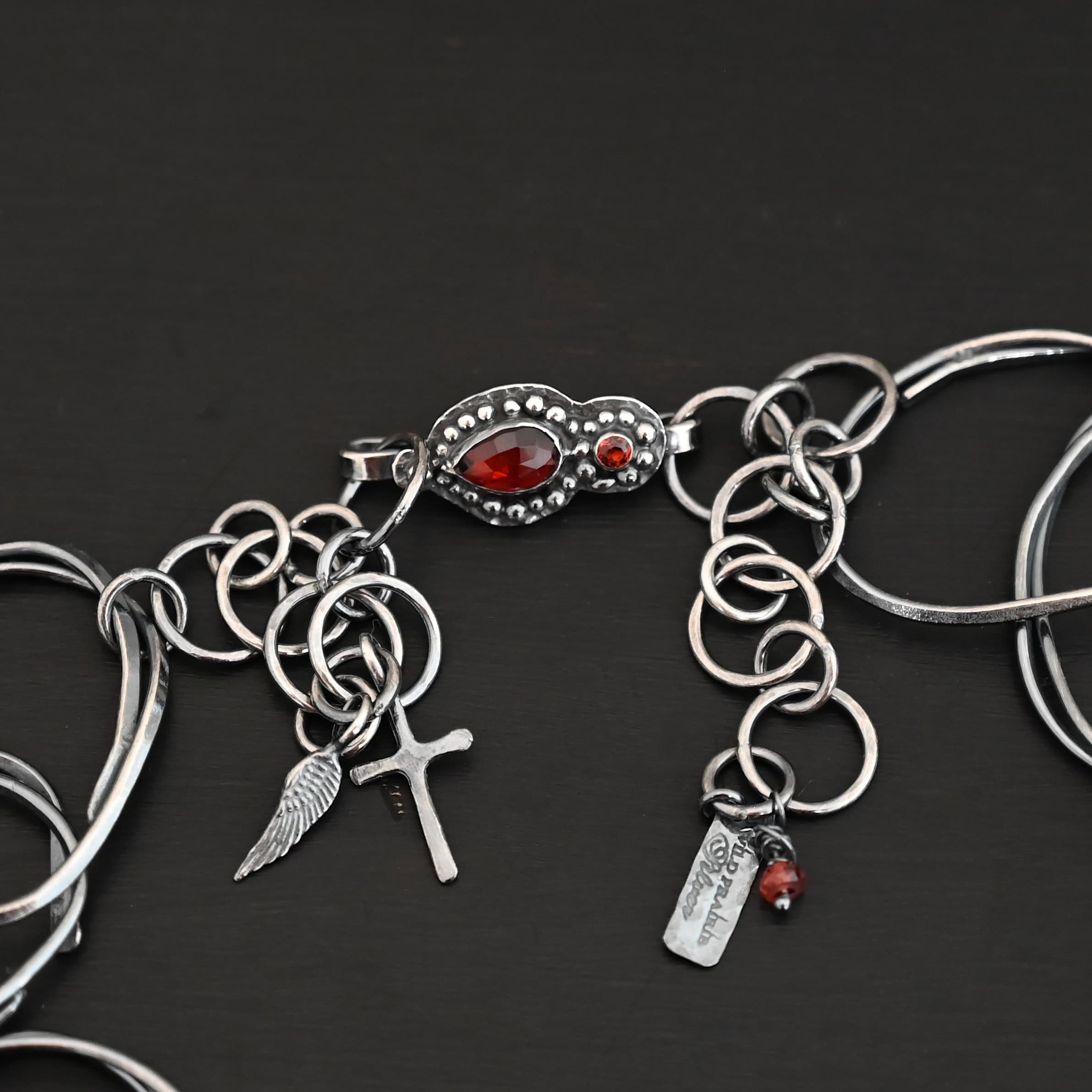 Clasp for the "Back To Our Roots Necklace." There are two garnets. one is a pear shape and the other is circular. Both have sterling silver ball detail around them. There is a small cross and angel wing charm attached to the silver extension.