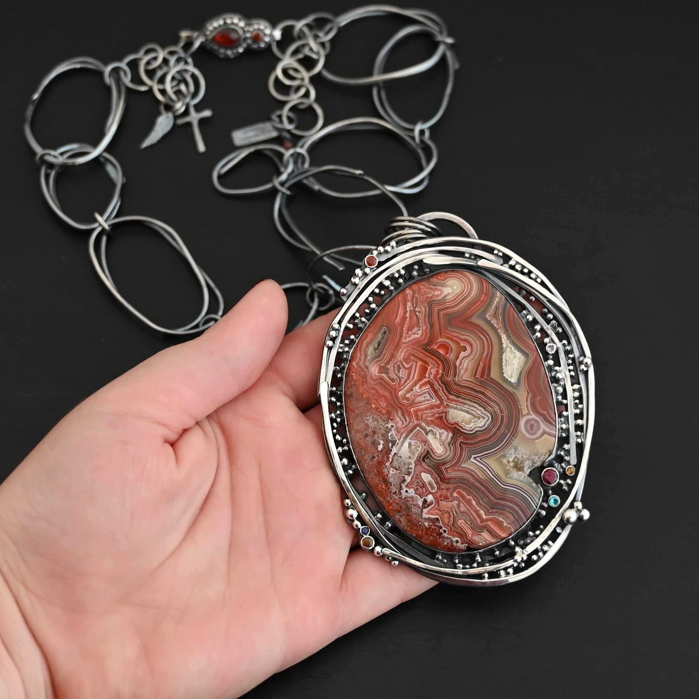 "Back To Our Roots" Necklace. There is a very large oval crazy lace agate in the center with multiple lines of sterling silver wrapped around it. There are small stones scattered around the pendant, along with lots of sterling silver ball detail. Chain is handmade out of large, sterling silver oval links that detach from the pendant to change the chain. Pendant is held in the fingertips of a hand.