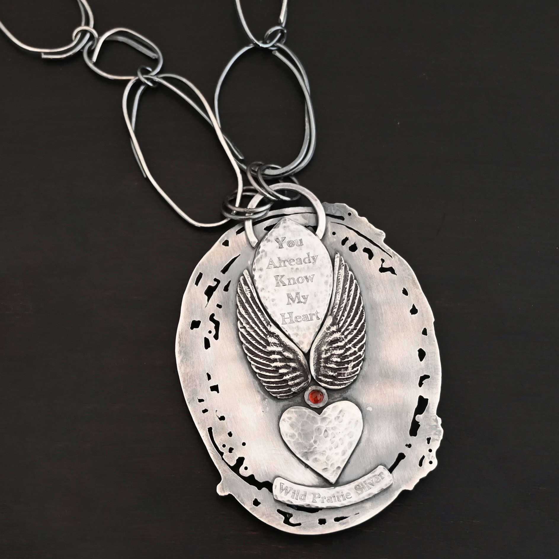 Back of the "Back To Our Roots" Necklace. There is a loop at the top and a marquise shape beneath it with the words, "You already know my heart" stamped into it. There is an angel wing on each side of the marquise shape. There is a small garnet in between the angel wings on the bottom. Beneath this is a hammered heart. Beneath that is a curved piece that says, "Wild Prairie Silver."